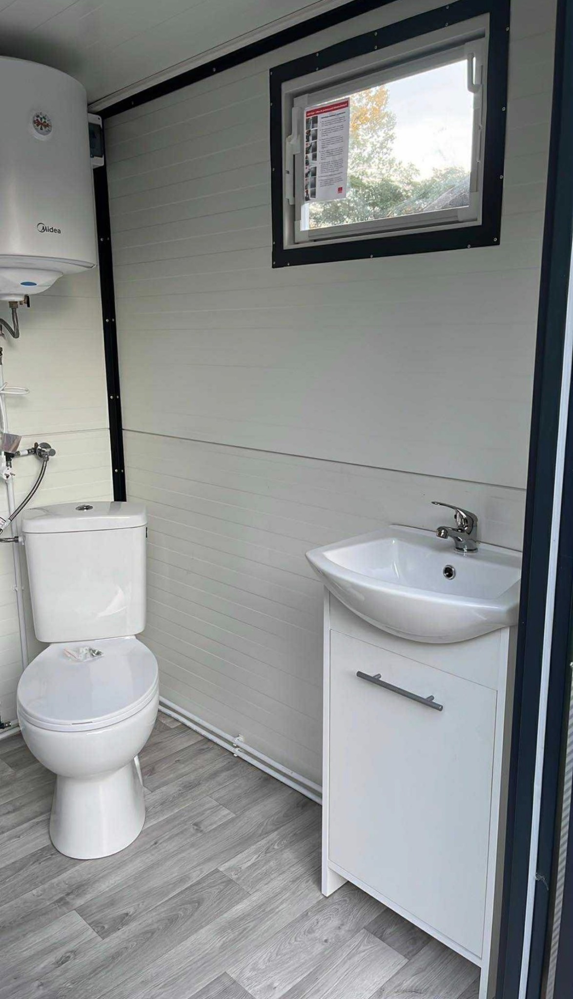 Nowoczesna, biała łazienka w kontenerze sanitarnym: WC, umywalka z szafką, bojler. Laminowane panele ścienne i podłogowe imitujące drewno. Okno z widokiem na zieleń.