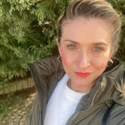 Portret kobiety z zielonymi oczami i blond włosami na tle zielonego krzewu, ubrana w biały sweter i zieloną kurtkę. Uśmiech, złote kolczyki.