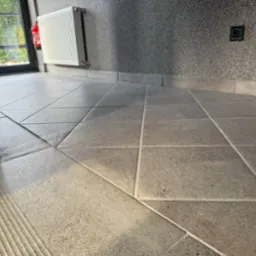 Wyczyszczona, szara, kwadratowa podłoga z płytek w budynku w Sosnowcu. Widoczny fragment schodów, grzejnik i gaśnica na ścianie. Świeżo po sprzątaniu.