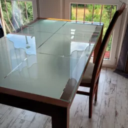 Stół z szklanym blatem i krzesłem na balkonie, widok na zieleń za oknem, jasne drewniane panele podłogowe, promienie światła na blacie.