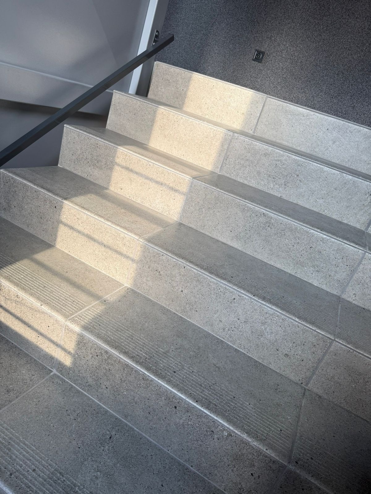 Schody wyłożone szarymi płytkami, z widocznymi promieniami słońca i czarną poręczą. Na podłodze widoczna szara wykładzina.