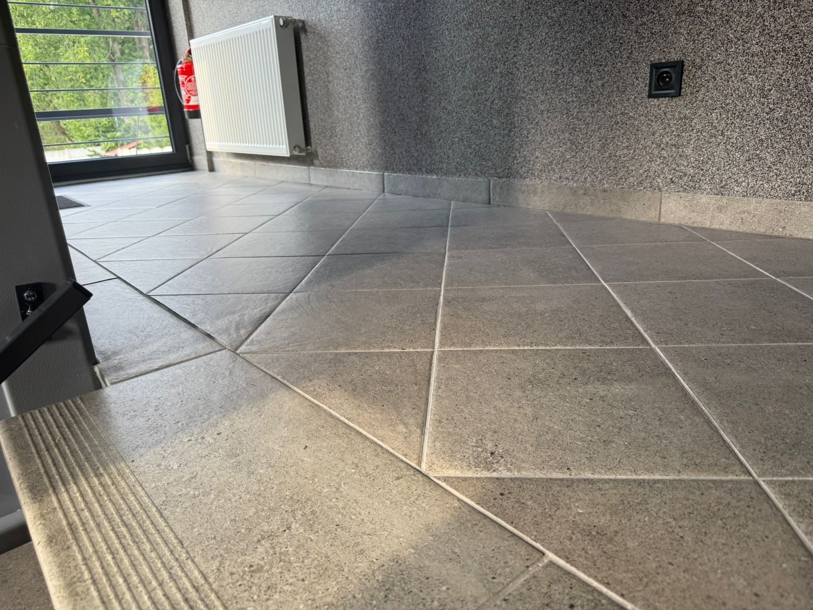 Wyczyszczona, szara, kwadratowa podłoga z płytek w budynku w Sosnowcu. Widoczny fragment schodów, grzejnik i gaśnica na ścianie. Świeżo po sprzątaniu.