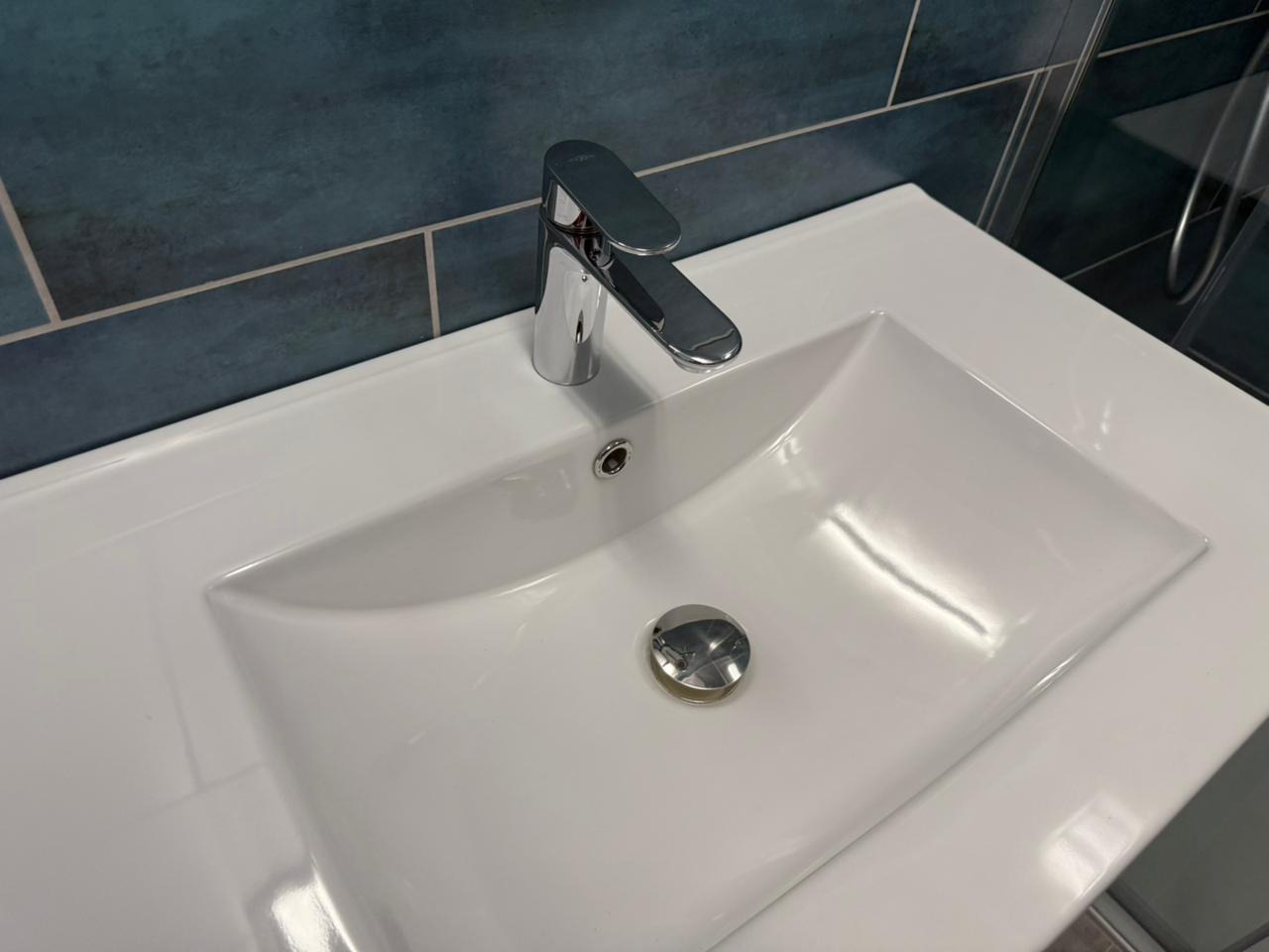 Biała umywalka z chromowaną baterią na tle niebieskich płytek. Widoczny odpływ i fragment kabiny prysznicowej. Czysta powierzchnia.