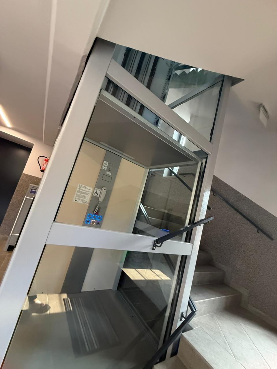 Wnętrze szybu windowego z kabiną dla niepełnosprawnych, widok z dołu na schody i fragment klatki schodowej. Widoczne panele sterowania i elementy konstrukcyjne.