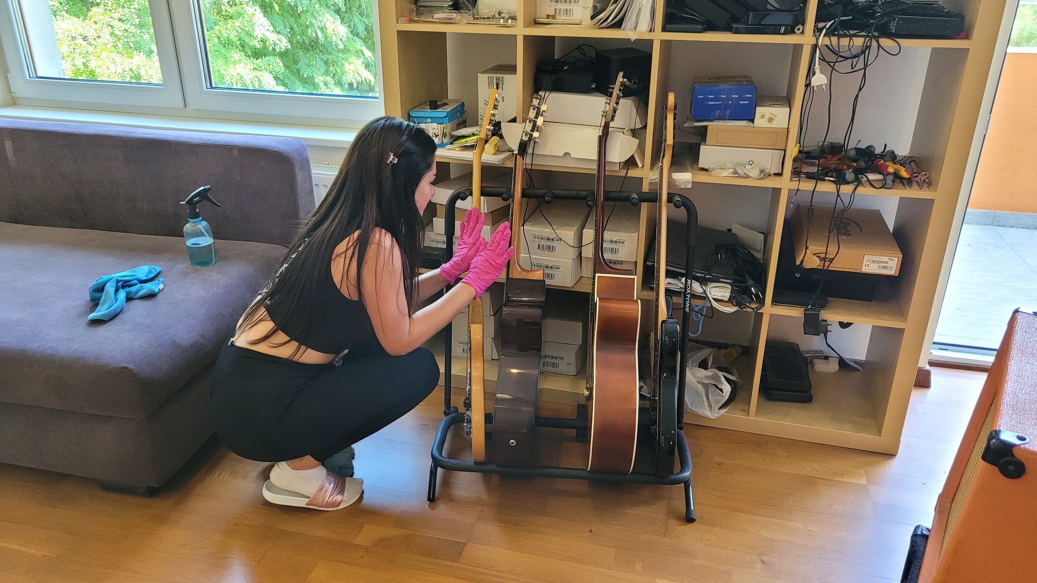 Kobieta w różowych rękawicach czyści gitary na stojaku w pokoju. W tle okno, sofa, szafka z różnymi przedmiotami. Widoczny płyn do czyszczenia i ściereczka.