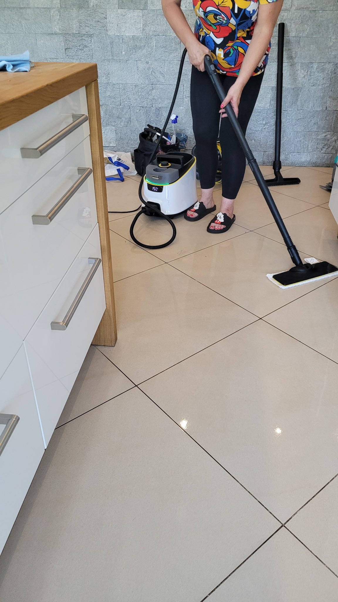 Osoba czyści podłogę mopem parowym Karcher, widoczny sprzęt i akcesoria. Beżowe płytki podłogowe i białe szafki z drewnianym blatem. Ujęcie z bliska.