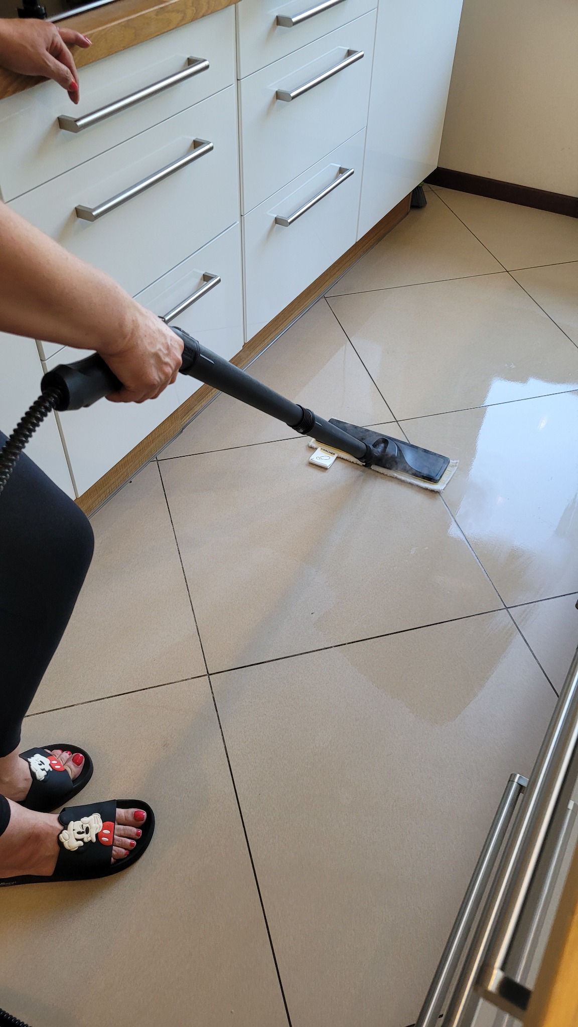 Czyszczenie podłogi parownicą Karcher w kuchni z białymi szafkami. Widoczne płytki podłogowe i fragment nóg osoby w klapkach z motywem Myszki Miki.
