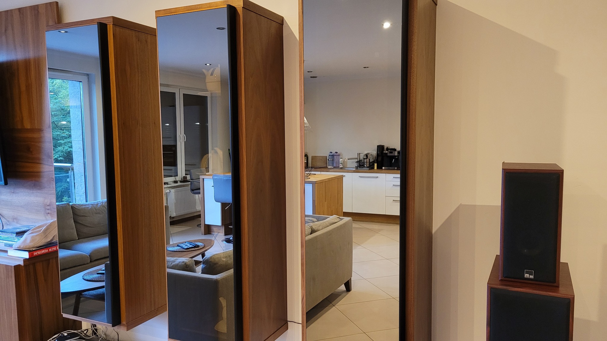 Wnętrze mieszkania z lustrami w drewnianych ramach odbijającymi salon i kuchnię, ukazując przestronność i jasność pomieszczeń. Widoczne meble i sprzęty.