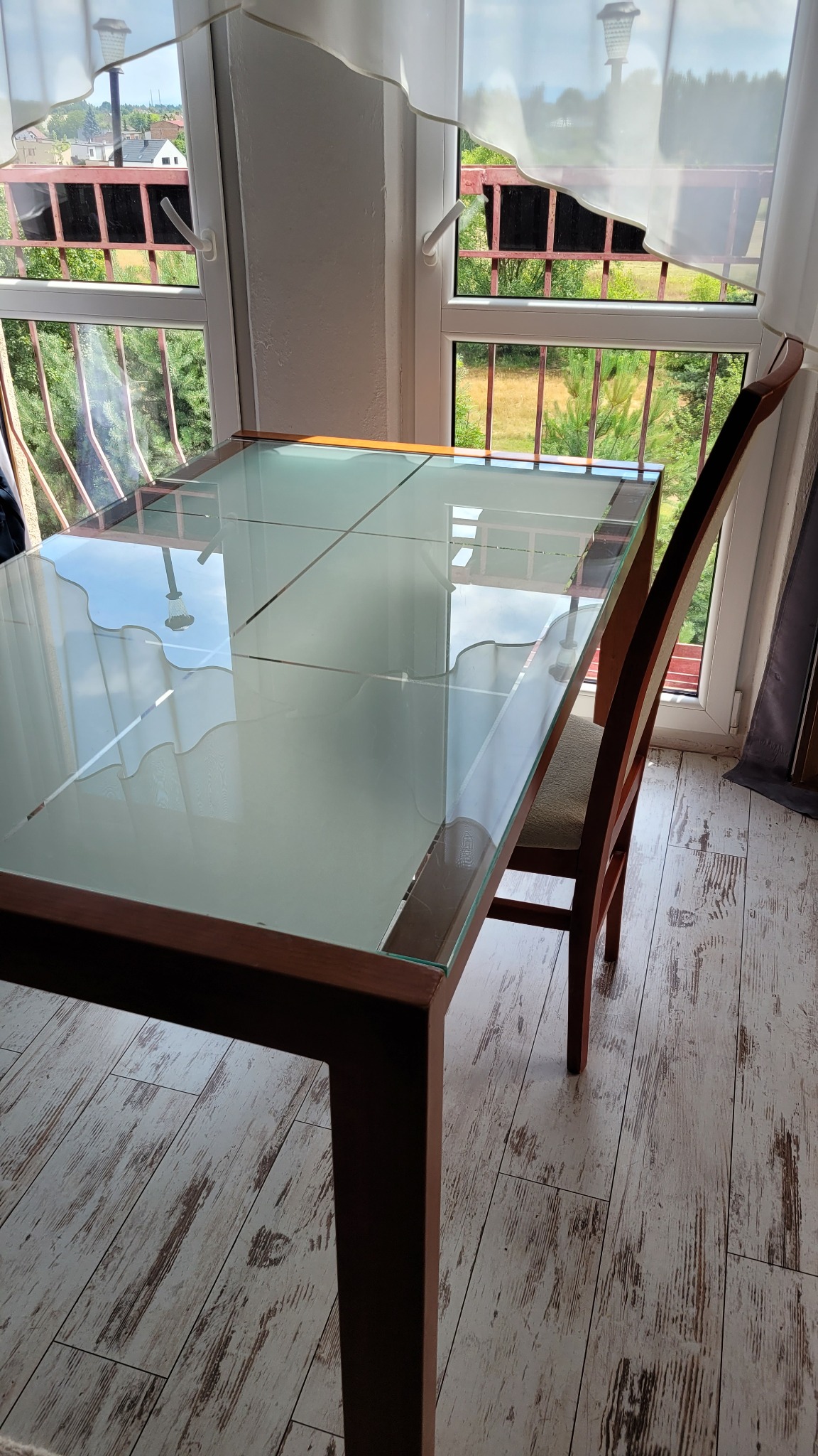 Stół z szklanym blatem i krzesłem na balkonie, widok na zieleń za oknem, jasne drewniane panele podłogowe, promienie światła na blacie.