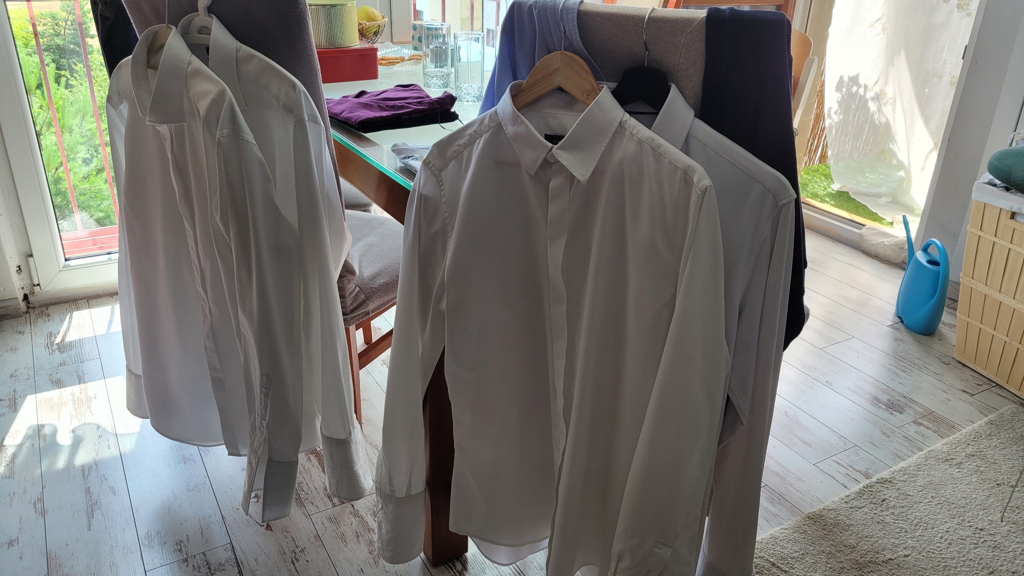 Wyprane, białe koszule wiszą na krzesłach w jasnym pokoju, czekając na prasowanie. Widoczna drewniana podłoga i elementy wyposażenia wnętrza.