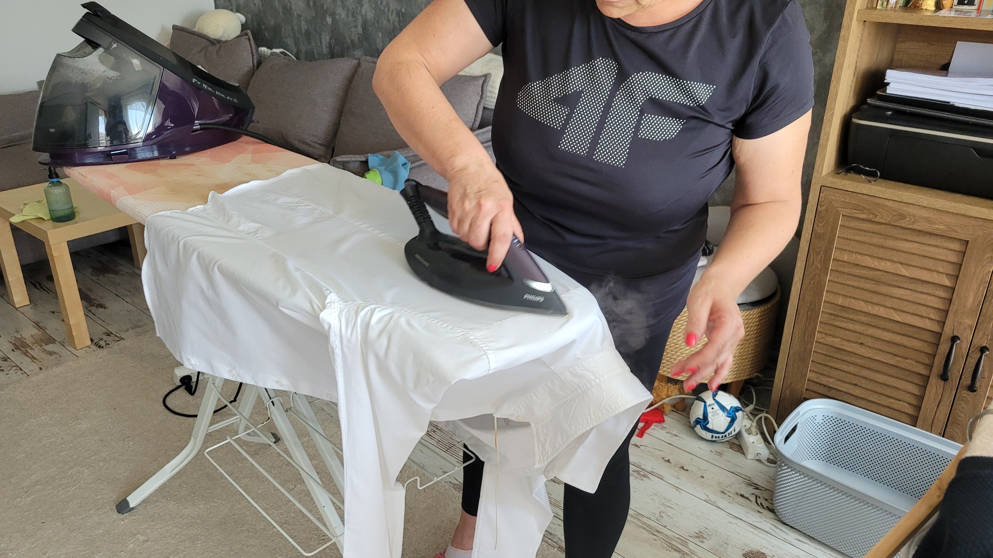Osoba prasuje białą koszulę parownicą Philips na desce do prasowania. W tle widoczny mebel i sofa. Prasowanie z użyciem pary.