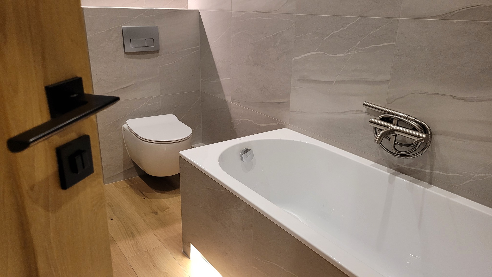 Elegancka łazienka po remoncie: biała wanna i podwieszana toaleta na tle szarych płytek imitujących marmur. Widoczna armatura i drewniane drzwi z czarną klamką.