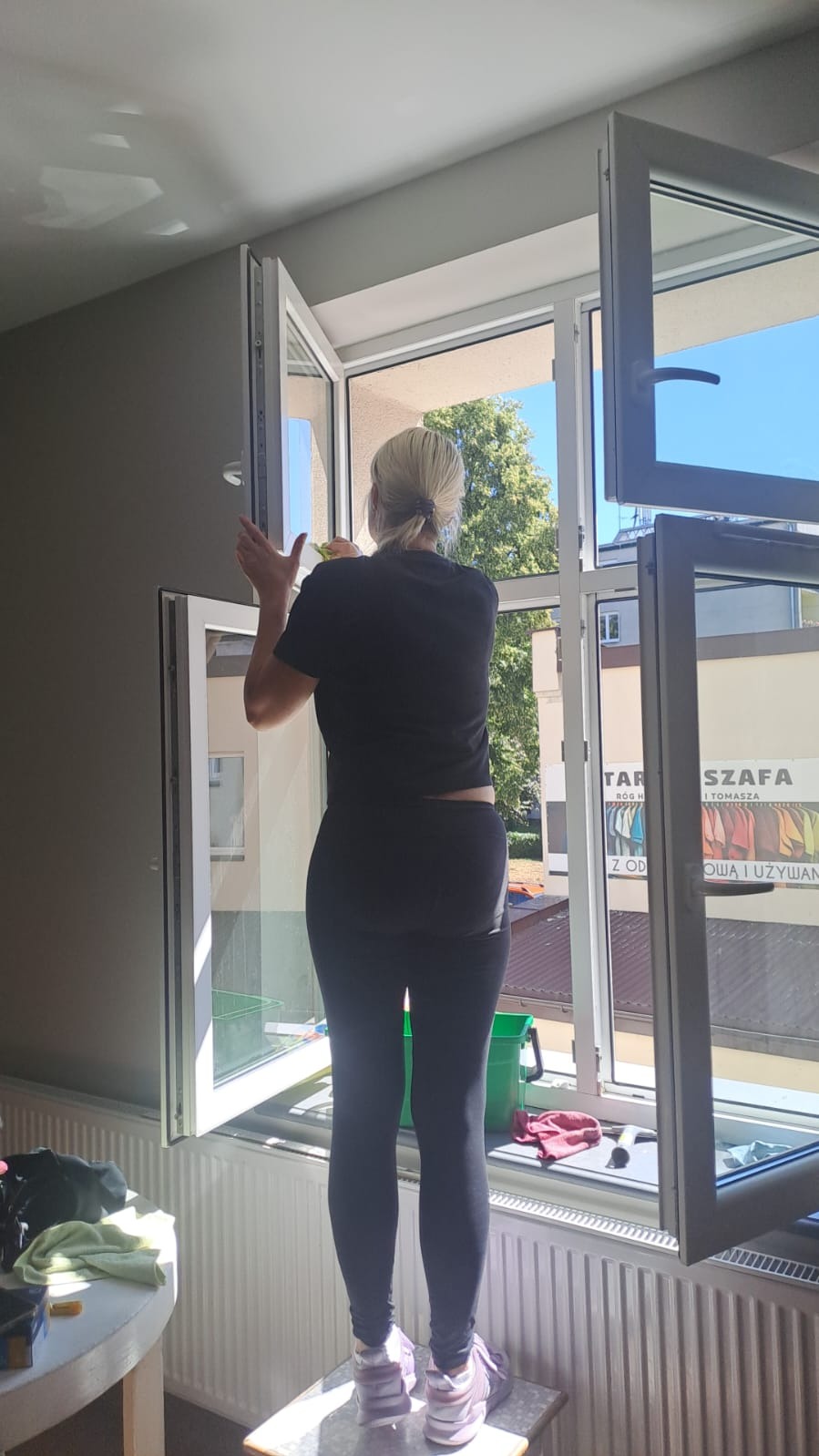 Kobieta w czarnych legginsach czyści okno w mieszkaniu, stojąc na stołku. Widoczne otwarte okna, budynki za oknem i akcesoria do mycia okien.