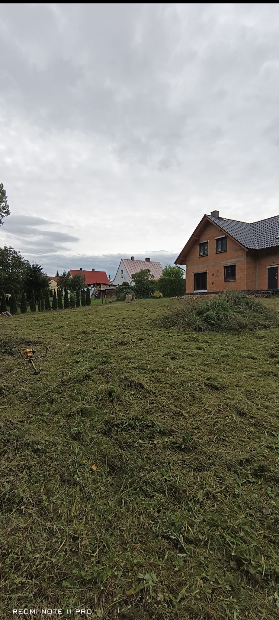 Po koszeniu: trawa skoszona na posesji z domem w budowie w tle. Na pierwszym planie leży kosa spalinowa.