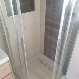 Daniel Daniluk - Nowoczesna kabina prysznicowa z jasnymi płytkami i wnęką na kosmetyki, wykończona drewnopodobną ścianą. Widoczny fragment szafki umywalkowej.