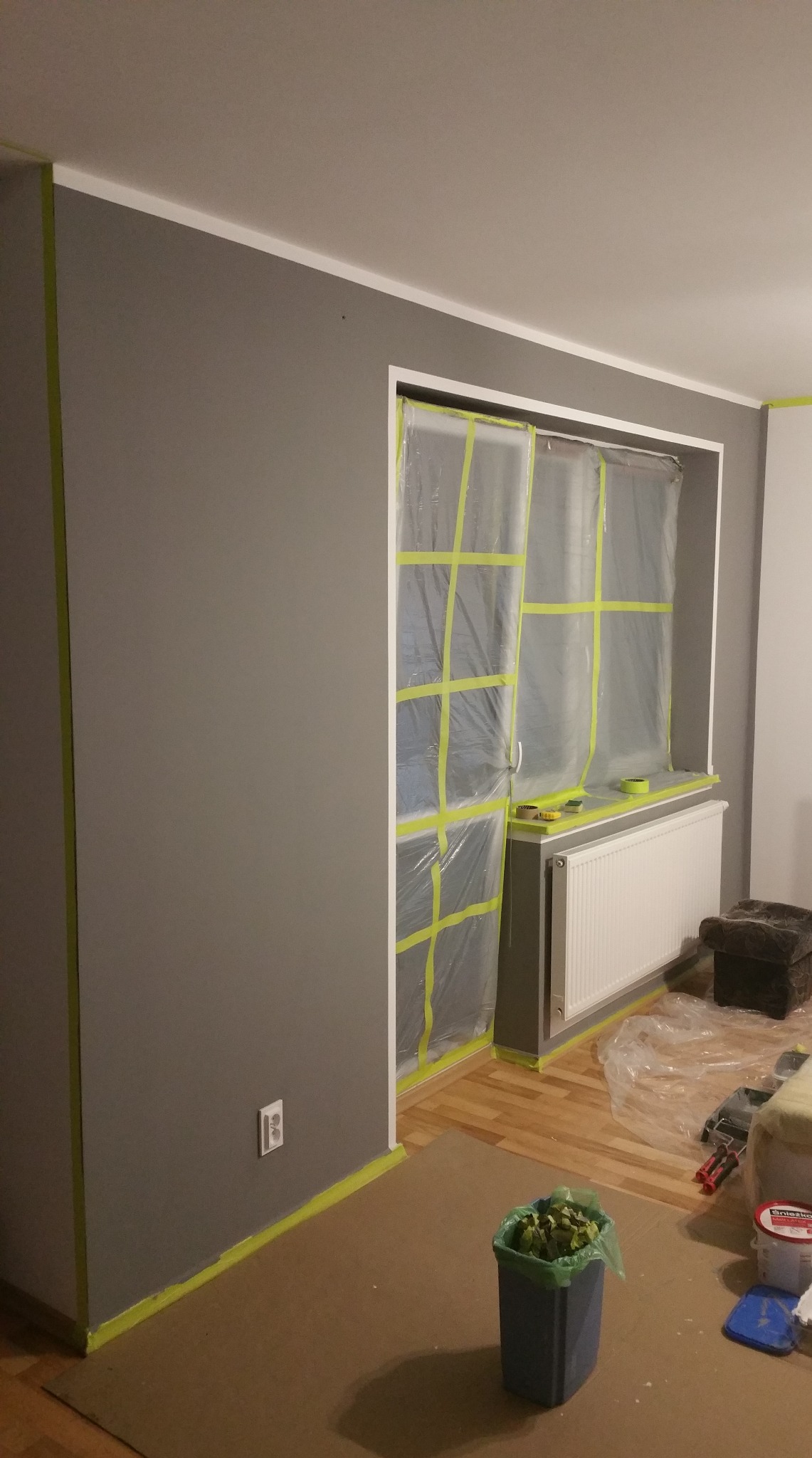 Pokój w trakcie malowania na szaro, okno zabezpieczone folią i taśmą, widoczny grzejnik i akcesoria malarskie. Podłoga zabezpieczona kartonem.