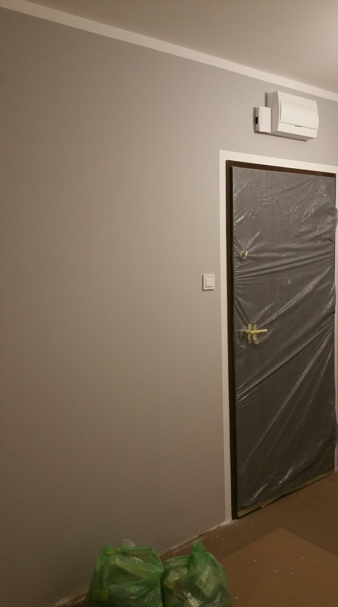 Ściana pomalowana na szaro, zabezpieczone drzwi folią malarską, widoczne worki z odpadami. Przygotowanie do malowania ścian w pomieszczeniu.