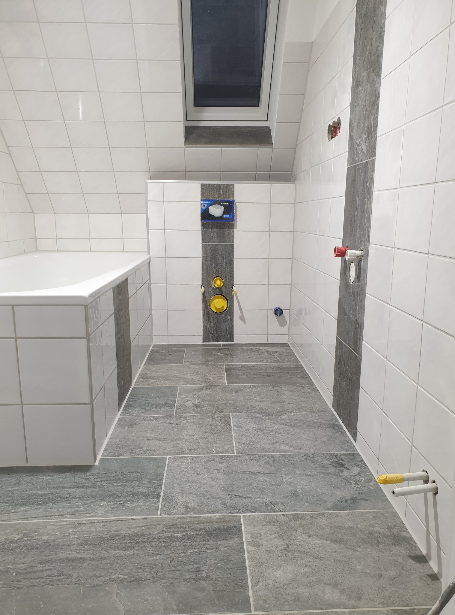 Wykończona łazienka na poddaszu z szarymi płytkami podłogowymi i białymi ściennymi. Widoczne przyłącza wodne i okno dachowe.