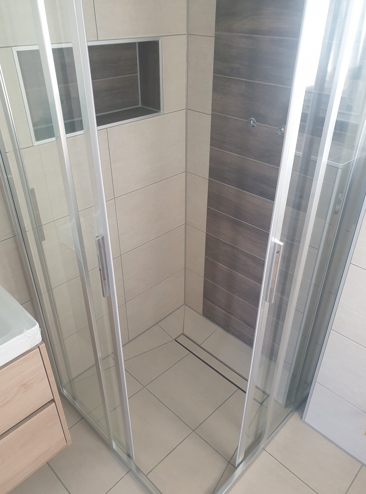 Nowoczesna kabina prysznicowa z jasnymi płytkami i wnęką na kosmetyki, wykończona drewnopodobną ścianą. Widoczny fragment szafki umywalkowej.