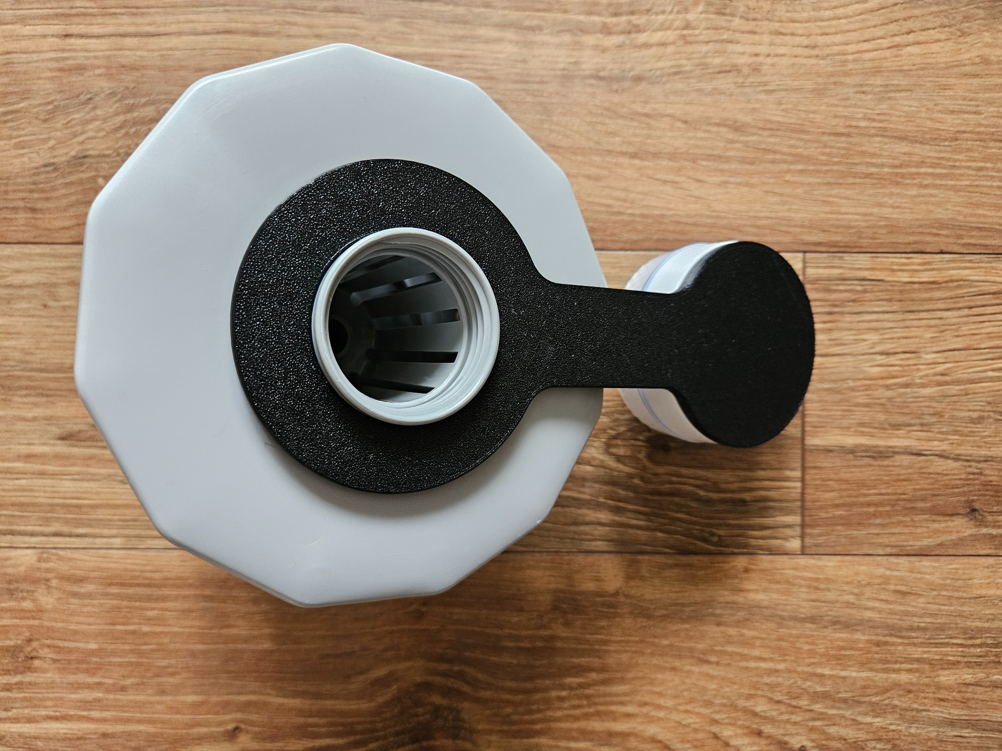 Szary, wielokątny pojemnik z czarnym uchwytem i białym gwintem na drewnianym podłożu, widok z góry, ukazujący detale wykonania i precyzję kształtu.