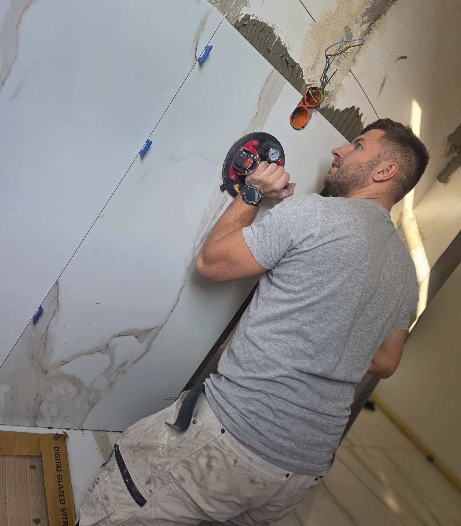 Mężczyzna w szarej koszulce montuje wielkoformatowe płytki z marmurowym wzorem na ścianie, używając przyssawki. Widoczne nieotynkowane fragmenty ściany i wystające kable.