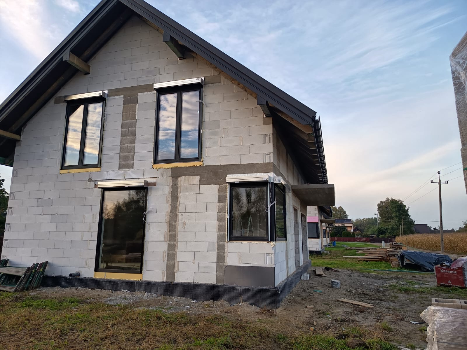 Surowy dom z betonu komórkowego z zamontowanymi oknami i częściowo opuszczonymi roletami zewnętrznymi, widok z boku na tle pochmurnego nieba.