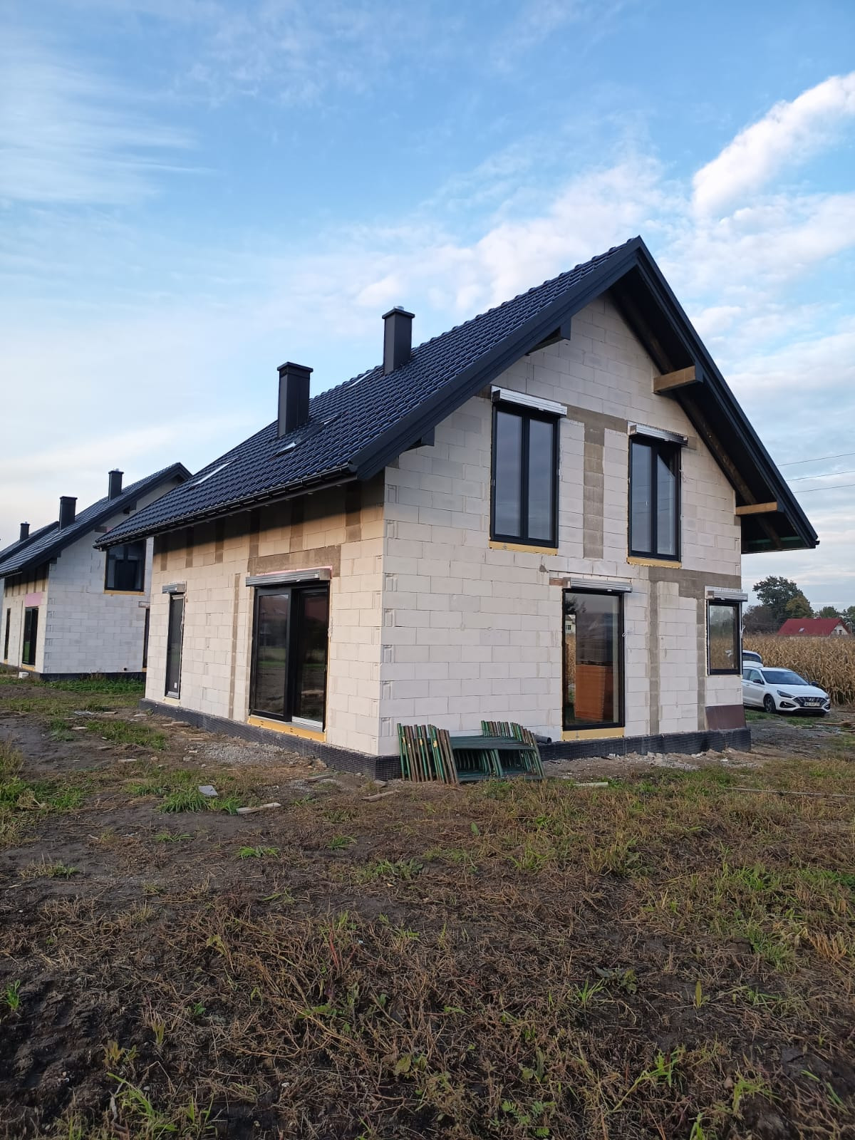 Dom w trakcie budowy z czarnymi oknami i roletami zewnętrznymi, widok z zewnątrz na surową elewację i dach z kominem, w tle drugi budynek i auto.