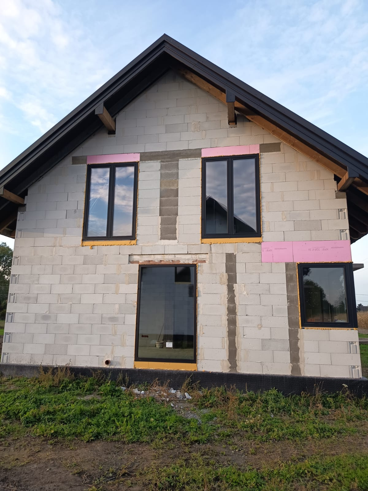 Montaż okien z czarnymi ramami w domu z białego bloczka, Leńcze. Widoczne różowe ocieplenie wokół okien. Okna odbijają niebo, budynek w stanie surowym.