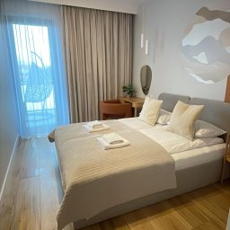 VADYM FEDORCHUK FIRMA USŁUGOWA - Jasna sypialnia z podwójnym łóżkiem, drewnianą podłogą i widokiem na balkon z krzesłem wiszącym. Minimalistyczny design, beżowa kolorystyka, spokojna atmosfera.