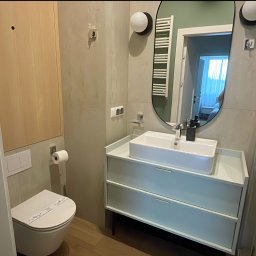 VADYM FEDORCHUK FIRMA USŁUGOWA - Nowoczesna, czysta łazienka po sprzątaniu w Kołobrzegu. Biała umywalka, toaleta z naklejką higieniczną, kabina prysznicowa i okrągłe lustro.