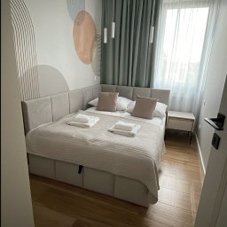 VADYM FEDORCHUK FIRMA USŁUGOWA - Jasna sypialnia z łóżkiem typu king-size, ozdobną tapetą i drewnianą podłogą. Widok przez otwarte drzwi. Minimalistyczny wystrój wnętrza.