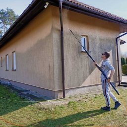 VADYM FEDORCHUK FIRMA USŁUGOWA - Mężczyzna w okularach myje elewację domu za pomocą teleskopowej lancy. Widoczny fragment elewacji, okno, trawnik i drzewa w tle. Jasny dzień.