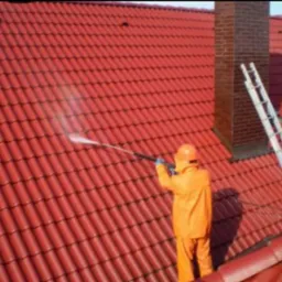 Czyszczenie dachu z czerwonej dachówki za pomocą myjki ciśnieniowej. Pracownik w pomarańczowym kombinezonie i kasku. Obok komin i drabina.