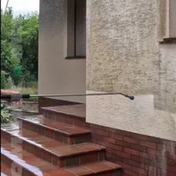 Czyszczenie elewacji domu myjką ciśnieniową, widoczne schody wejściowe z płytek i fragment ściany z efektem przed i po czyszczeniu. Świeżo umyte schody.