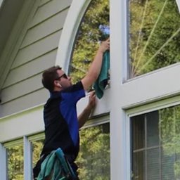 VADYM FEDORCHUK FIRMA USŁUGOWA - Pracownik w Kołobrzegu myje okno o nietypowym, łukowym kształcie, używając ściereczki. Widoczne drzewa odbijające się w szybie i narzędzia przy pasie.