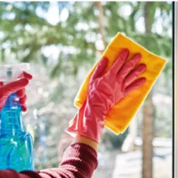 Osoba w różowych rękawicach czyści okno żółtą ściereczką i niebieskim płynem do mycia okien. Widok na zewnątrz przez czyszczone okno.