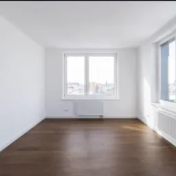 Jasne, puste wnętrze z drewnianą podłogą i białymi ścianami. Dwa okna z widokiem na miasto w tle. Minimalistyczny design, czyste linie i dużo naturalnego światła.