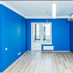 VADYM FEDORCHUK FIRMA USŁUGOWA - Pomalowane na niebiesko wnętrze pokoju z białymi drzwiami balkonowymi, grzejnikiem i klimatyzatorem. Podłoga wyłożona panelami w odcieniach szarości. Dwa cylindryczne białe klosze.