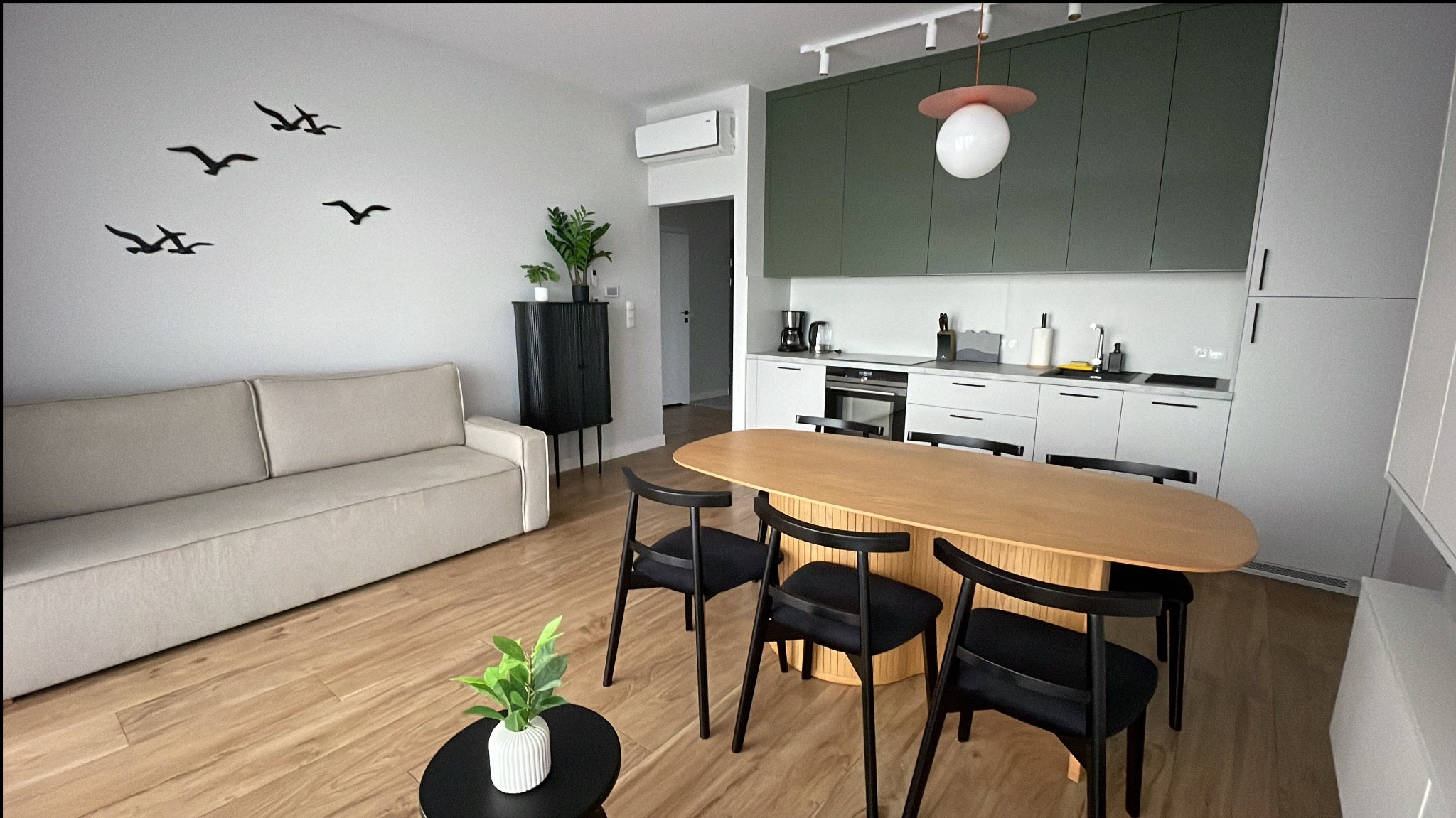Nowoczesne, czyste wnętrze mieszkania z drewnianym stołem, czarnymi krzesłami i minimalistyczną kuchnią w odcieniach bieli i zieleni. Na ścianie dekoracja z motywem ptaków.