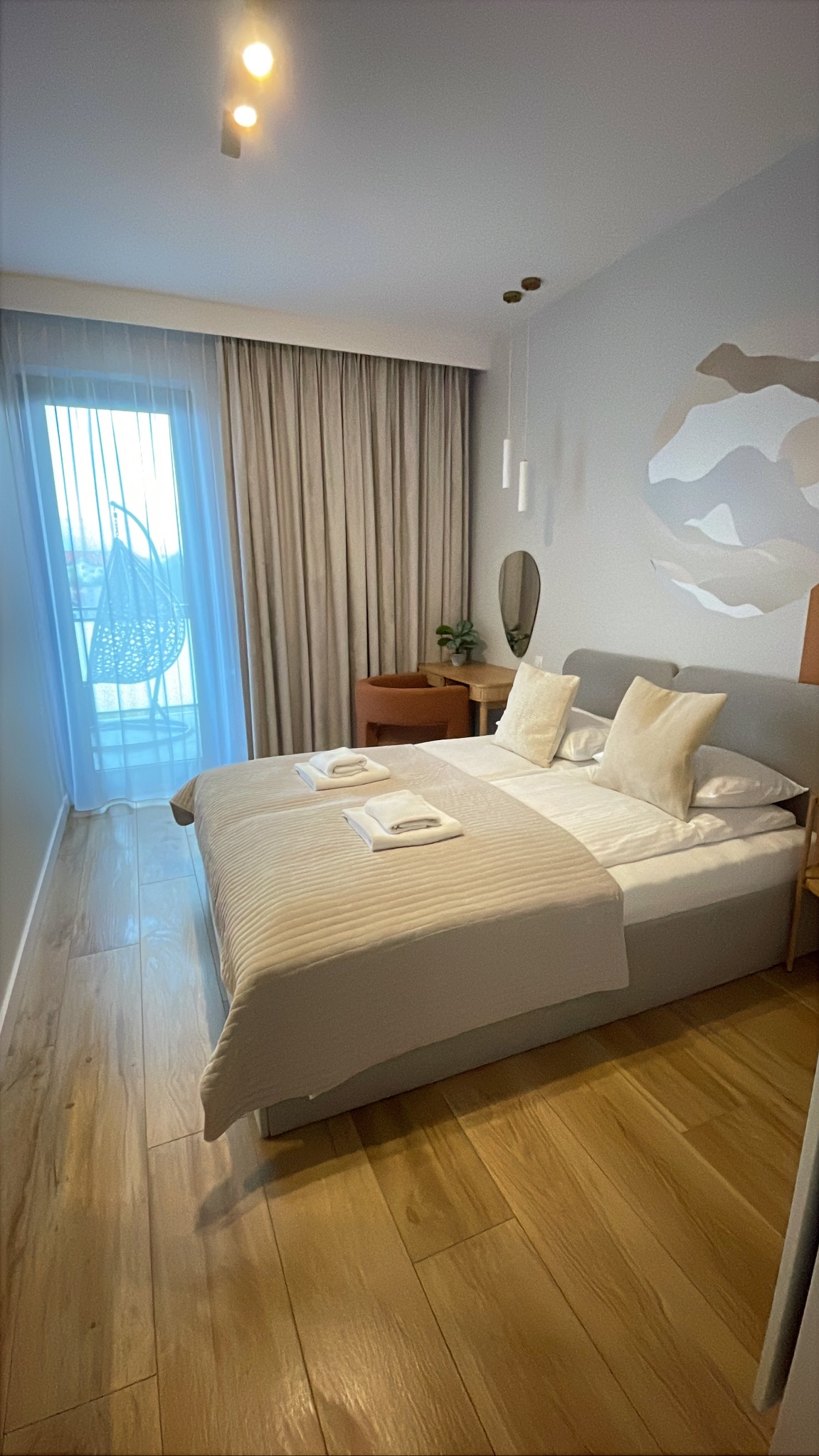 Jasna sypialnia z podwójnym łóżkiem, drewnianą podłogą i widokiem na balkon z krzesłem wiszącym. Minimalistyczny design, beżowa kolorystyka, spokojna atmosfera.