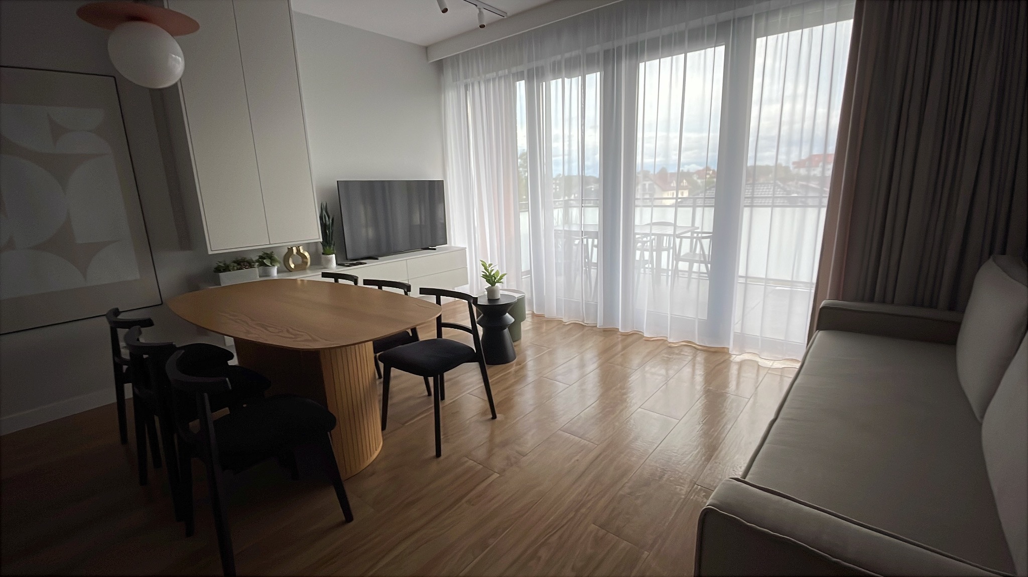 Jasne, przestronne wnętrze apartamentu w Kołobrzegu z drewnianym stołem i krzesłami, widokiem na balkon przez duże okna z białymi zasłonami.