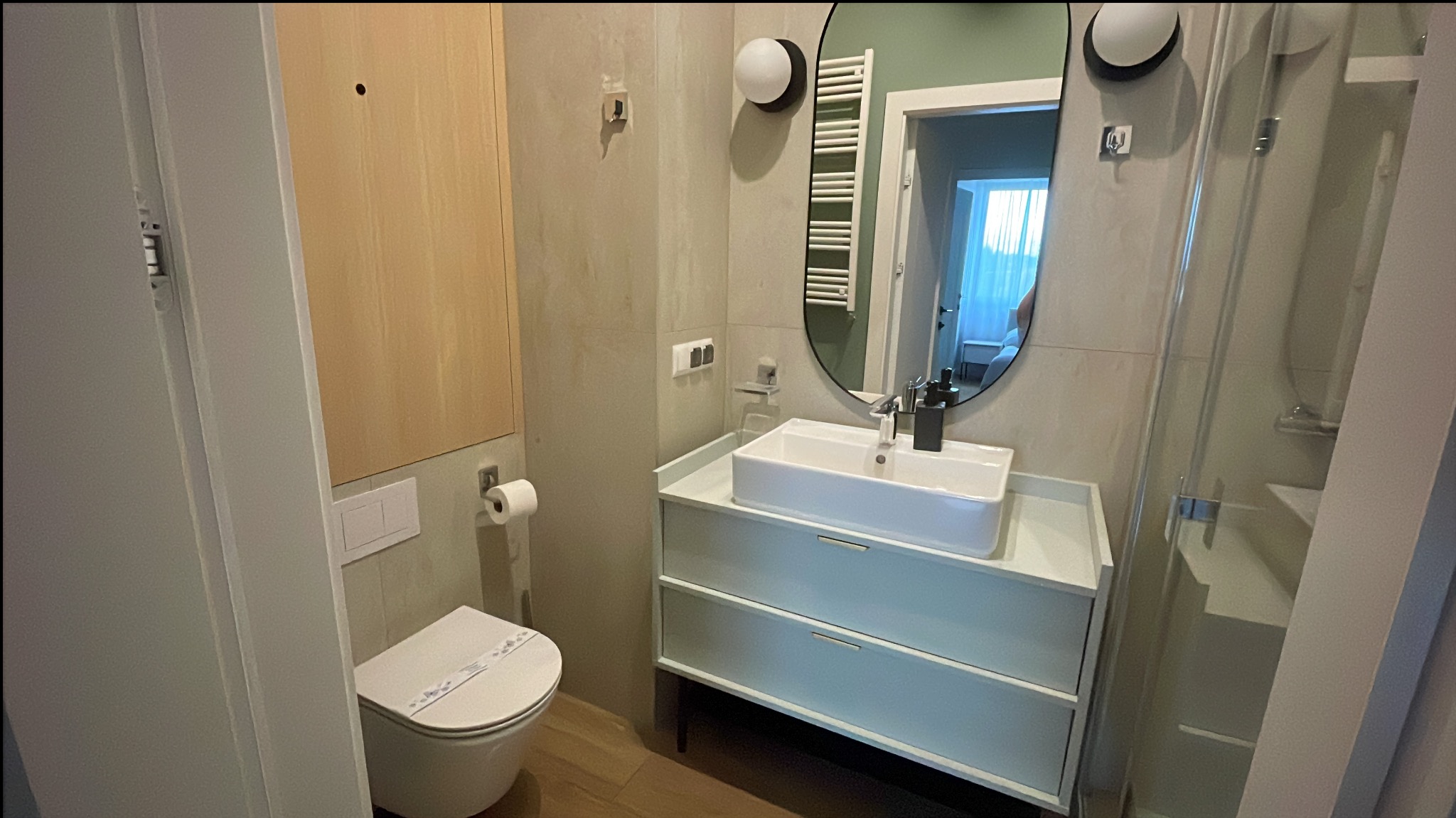 Nowoczesna, czysta łazienka po sprzątaniu w Kołobrzegu. Biała umywalka, toaleta z naklejką higieniczną, kabina prysznicowa i okrągłe lustro.