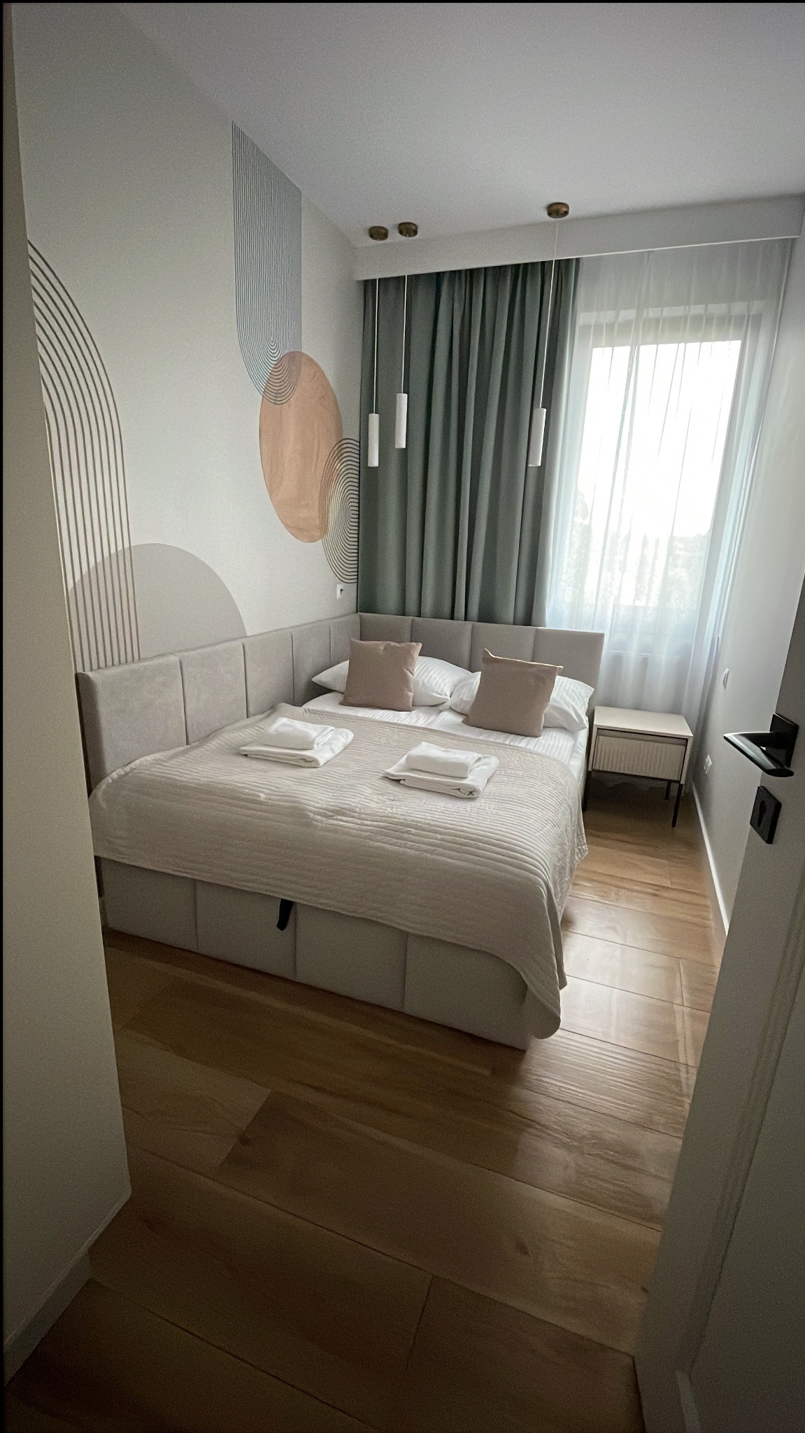 Jasna sypialnia z łóżkiem typu king-size, ozdobną tapetą i drewnianą podłogą. Widok przez otwarte drzwi. Minimalistyczny wystrój wnętrza.