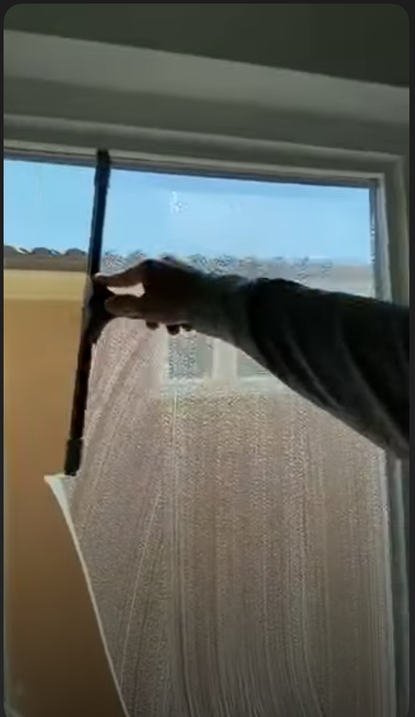 Mycie okna z użyciem ściągaczki. Widoczne smugi wody na szybie i fragment dłoni osoby wykonującej usługę. W tle jasne niebo i budynek.