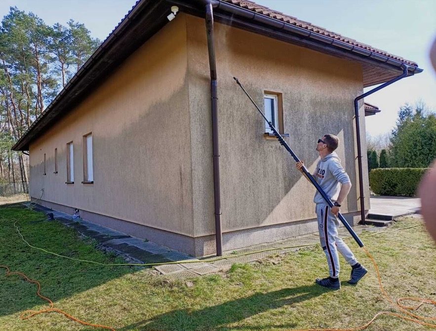 Mężczyzna w okularach myje elewację domu za pomocą teleskopowej lancy. Widoczny fragment elewacji, okno, trawnik i drzewa w tle. Jasny dzień.