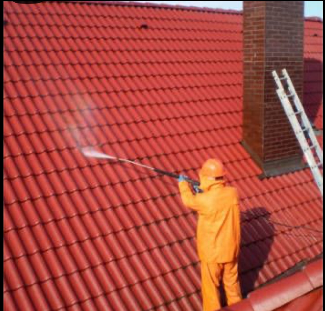 Czyszczenie dachu z czerwonej dachówki za pomocą myjki ciśnieniowej. Pracownik w pomarańczowym kombinezonie i kasku. Obok komin i drabina.