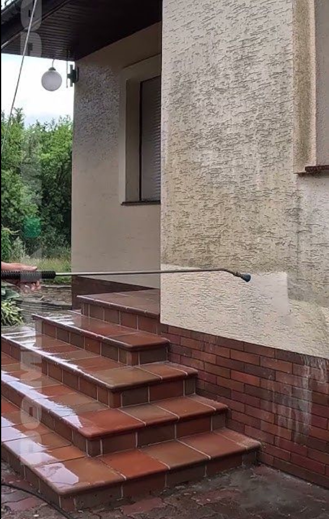 Czyszczenie elewacji domu myjką ciśnieniową, widoczne schody wejściowe z płytek i fragment ściany z efektem przed i po czyszczeniu. Świeżo umyte schody.