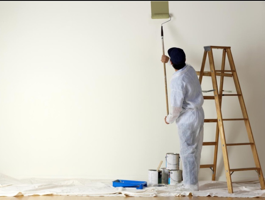 Malarz w kombinezonie maluje ścianę wałkiem na długim kiju. Obok drabina, puszki z farbą i kuweta. Widok z tyłu, jasne wnętrze.