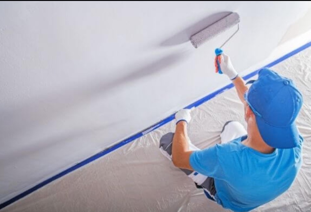 Malowanie sufitu wałkiem przez malarza w niebieskiej czapce. Zabezpieczona taśmą krawędź sufitu. Widok z góry na pracownika podczas pracy.