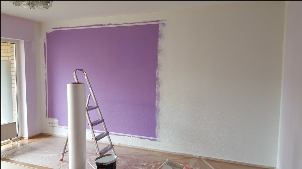 Ściana w trakcie malowania na fioletowo, z widoczną drabiną, wałkiem i folią ochronną na podłodze. Część ściany pomalowana, reszta biała. Widoczne niedociągnięcia przy suficie.